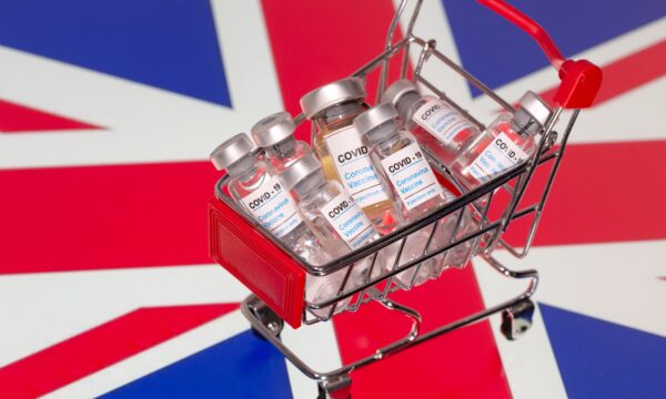 A small shopping basket filled with vials labeled "COVID-19 - Coronavirus Vaccine" is placed on a UK flag