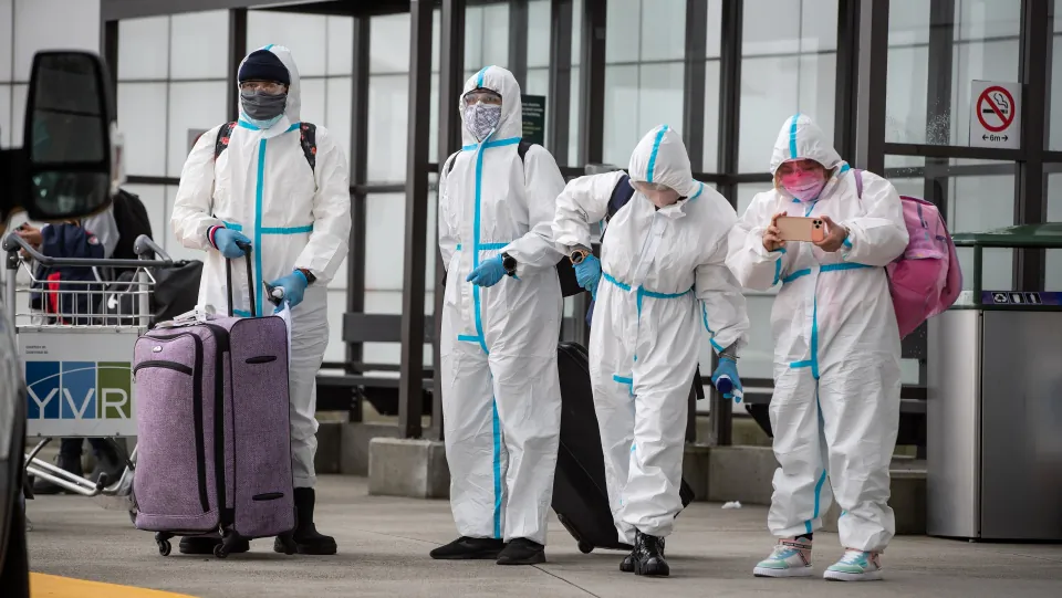 People wearing protective face masks, goggles and Tyvek suits who said they traveled from Colombia wait for a car rental company shuttle, after arriving at Vancouver International Airport in Richmond, B.C., on Dec. 31, 2020. (The Canadian Press/Darryl Dyck)