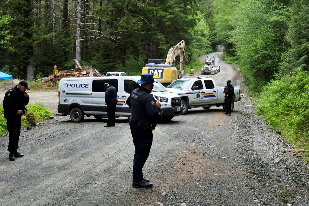 An excavator works in the background as police establish a checkpoint on McClure Main in the Caycuse area on B.C.'s Vancouver Island, on May 17, 2021. (Jen Osborne/The Canadian Press)