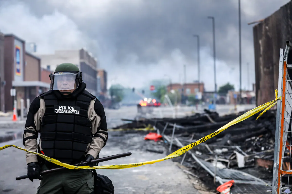 A police officer stands amid smoke and debris as buildings continue to burn in the aftermath of a night of protests and violence following the death of George Floyd in Minneapolis, Minn., on May 29, 2020. (Charlotte Cuthbertson/The Epoch Times)