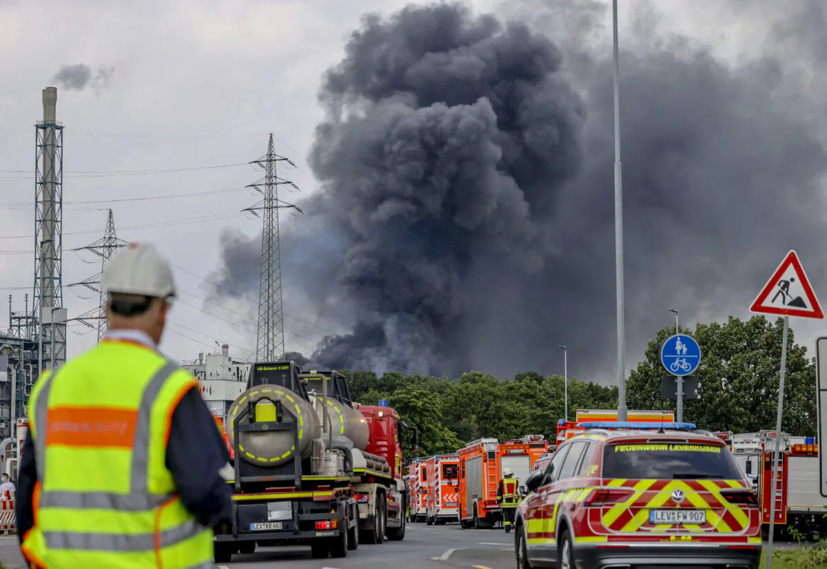Emergency vehicles of the fire brigade, rescue services, and police stand not far from an access road to the Chempark over which a dark cloud of smoke is rising in Leverkusen, Germany, on July 27, 2021. (Oliver Berg/dpa via AP)