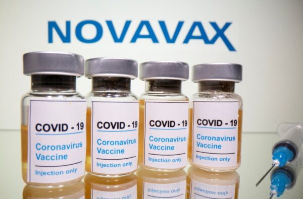 Vials and medical syringes are displayed in front of the Novavax logo in this figure