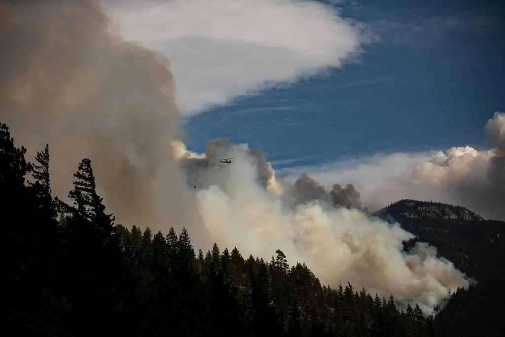 A helicopter carrying a water bucket flies past the Lytton Creek wildfire burning in the mountains near Lytton, B.C., on August 15, 2021. (The Canadian Press/Darryl Dyck)
