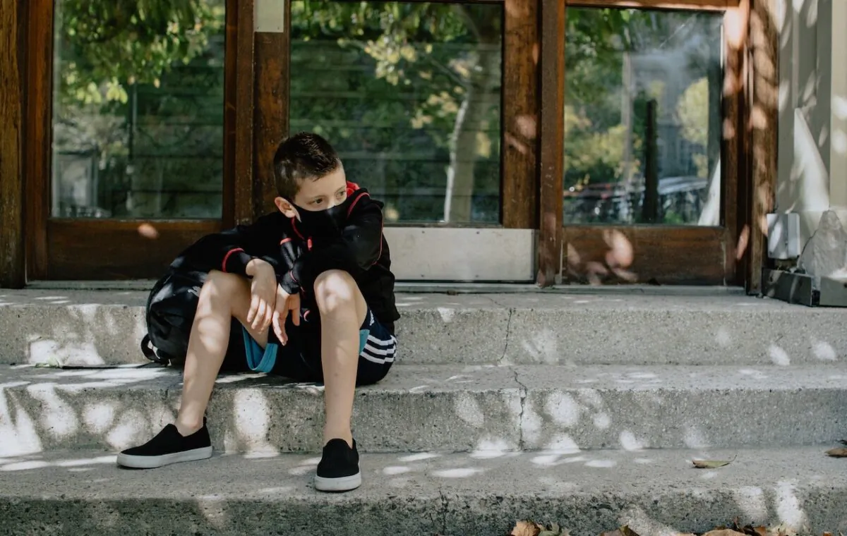 A boy wearing a mask sits on steps while waiting for school. (Kelly Sikkema/Unsplash)
