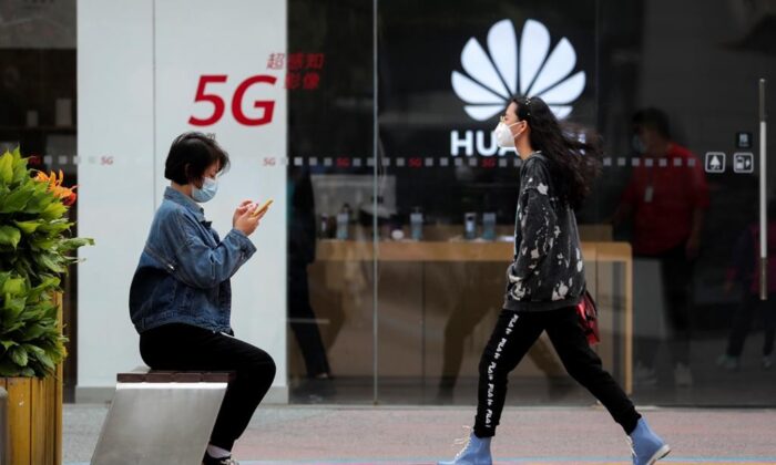 In this Oct. 11, 2020, file photo, a woman wearing a face mask to help curb the spread of the coronavirus browses her smartphone as a masked woman walks by the Huawei retail shop promoting it 5G network in Beijing.  Chinese tech giant Huawei said Wednesday, March 31, 2021, it eked out a gain in sales and profit last year but growth plunged after its smartphone unit was hammered by U.S. sanctions imposed in a fight with Beijing over technology and security. (AP Photo/Andy Wong, File)