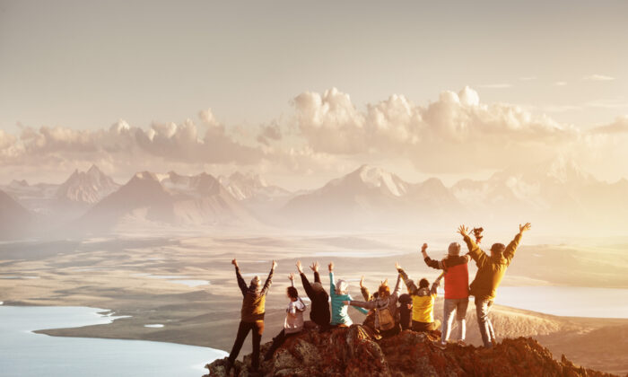 Big group of people having fun in success pose with raised arms on mountain top against sunset lakes and mountains. [Shutterstock]