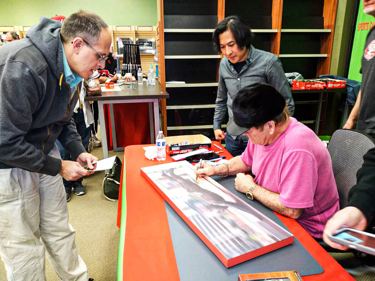 Pictured here with Baseball Legend Pete Rose is renowned sculptor Tom Tsuchiya (standing behind Rose) who sculpted the bronze statue of Rose outside Great American Ballpark in Cincinnati. Rose is signing a canvas print of the statue that depicts Rose in his trademark head-first slide, on Dec. 3, 2021. (Photo by Michael Sakal/The Epoch Times).
