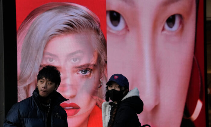 A woman wearing a mask stands near an ad featuring models for makeup products, in Beijing, on Dec. 28, 2021. (Ng Han Guan/AP Photo)