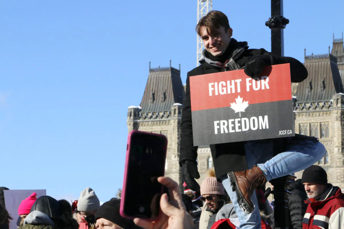 A man hangs from a lamp post and holds a placard from the Justice Centre for Constitutional Freedoms in front of Parliament Hill in Ottawa on Jan. 29, 2022. (Noé Chartier/The Epoch Times)