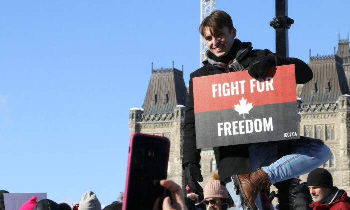 A man hangs from a lamp post and holds a placard from the Justice Centre for Constitutional Freedoms in front of Parliament Hill in Ottawa on Jan. 29, 2022. (Noé Chartier/The Epoch Times)