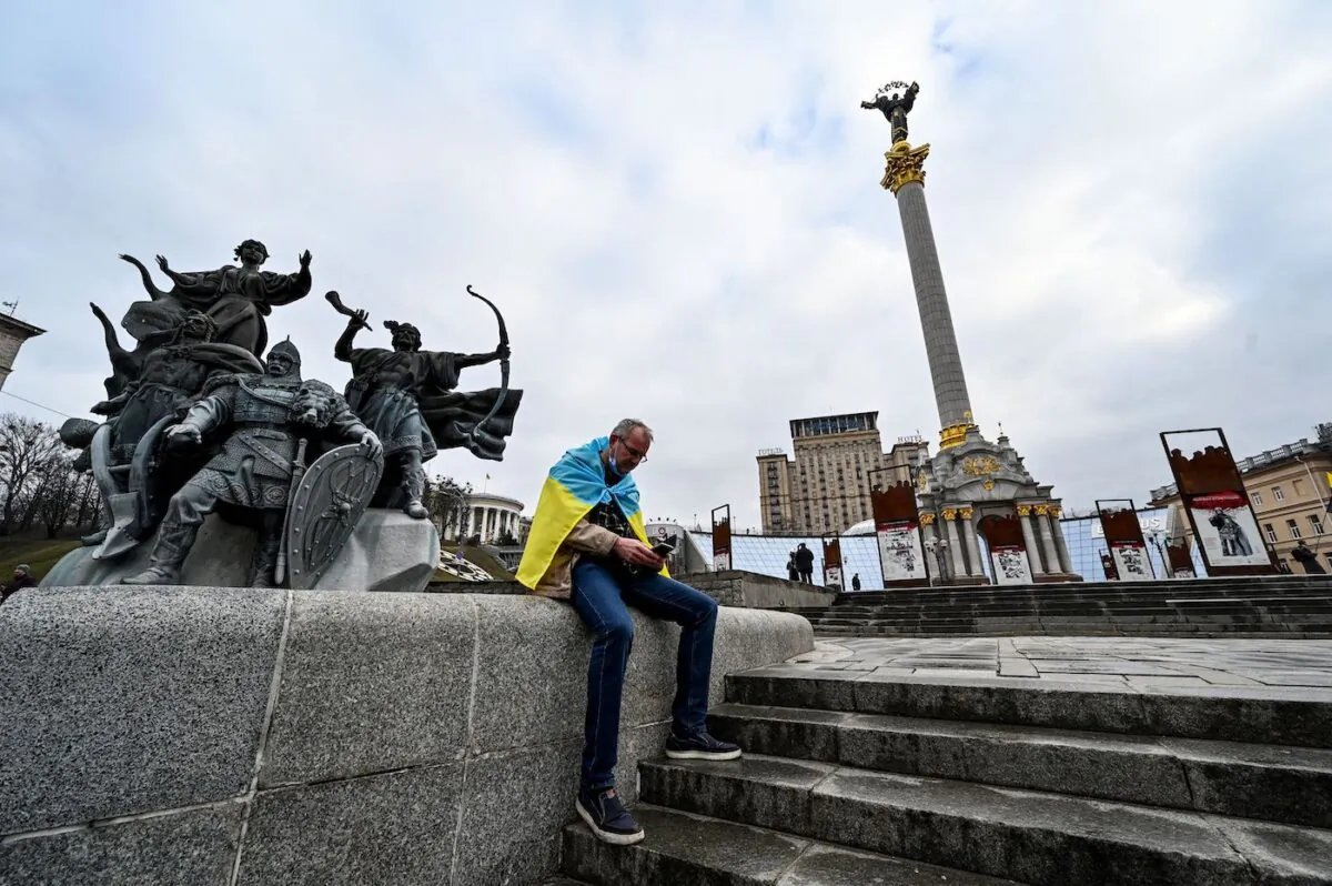 A man wrapped with a Ukrainian national flag watches news on his mobile phone as he sits at Maidan Independence Square in Kyiv, Ukraine, on Feb. 24, 2022. (Sergei Supinsky/AFP via Getty Images)