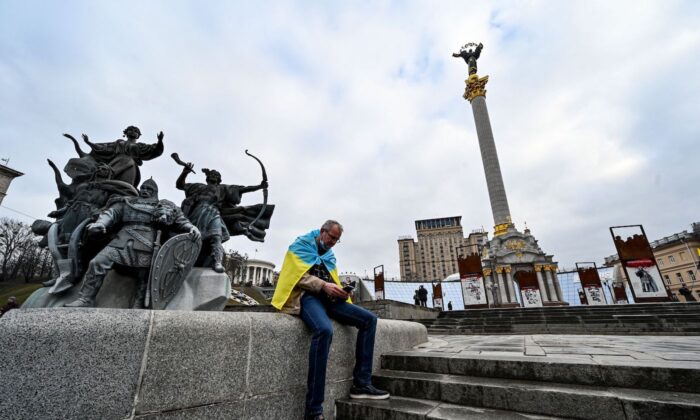 2022 年 2 月 24 日，一名裹着乌克兰国旗的男子坐在乌克兰基辅独立广场的手机上观看新闻。（Sergei Supinsky/AFP via Getty Images）