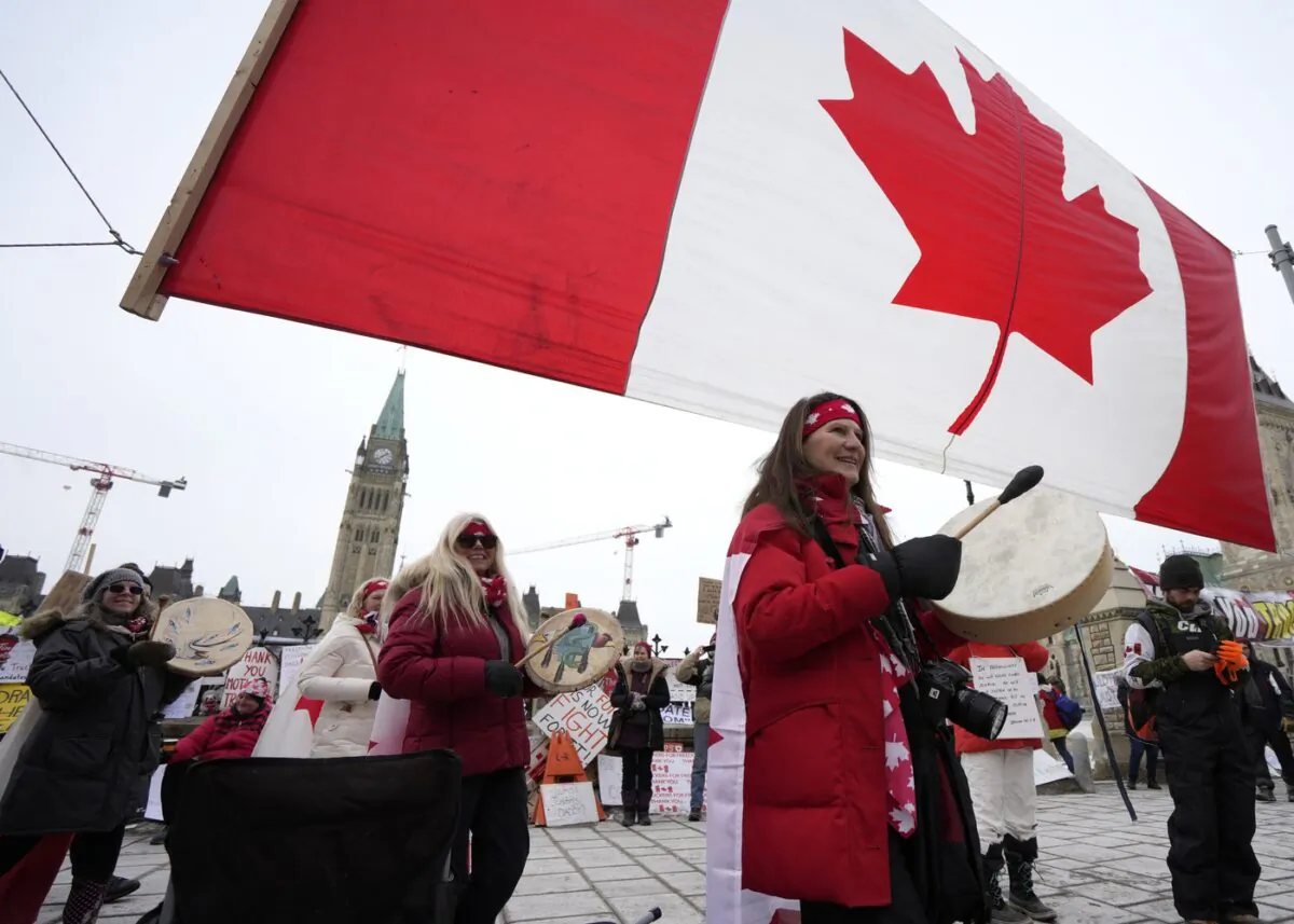 Women drum as they pass under a giant Canadian flag in front of Parliament Hill on the 20th day of a protest against COVID-19 measures that has grown into a broader anti-government protest, in Ottawa, on Feb. 16, 2022. (The Canadian Press/Justin Tang)