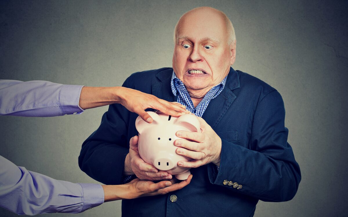 Elderly upset scared business man holding piggy bank trying to protect his savings from being stolen isolated gray background. (pathdoc/Shutterstock)