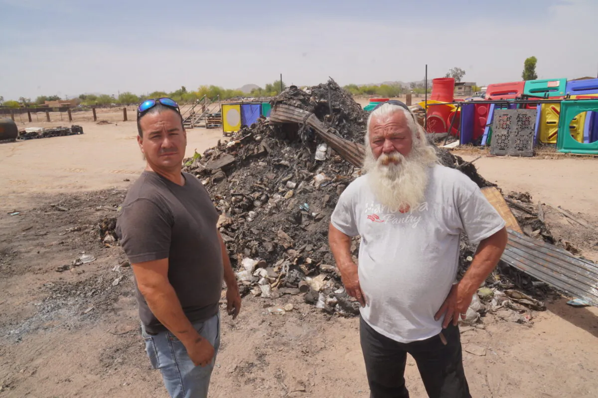 Tim Bennett, warehouse manager at the Maricopa Food Pantry in Maricopa, Ariz. (L), and pantry CEO Jim Shoaf stand in front of a pile of burned remains of six trailers and 48,000 pounds of food that went up in an electrical fire at the pantry on March 28, 2022. (Allan Stein/The Epoch Times)