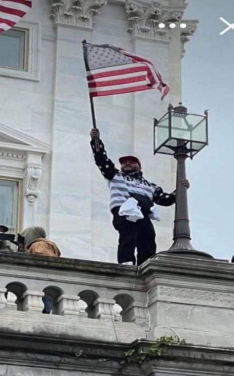 Screenshot of an image from Kash Kelly's Cell phone, showing Kash Lee Kelly standing on the ledge of the staircase at the Capitol Building on January 6, waving an American Flag, which was included in the Criminal Complaint. 