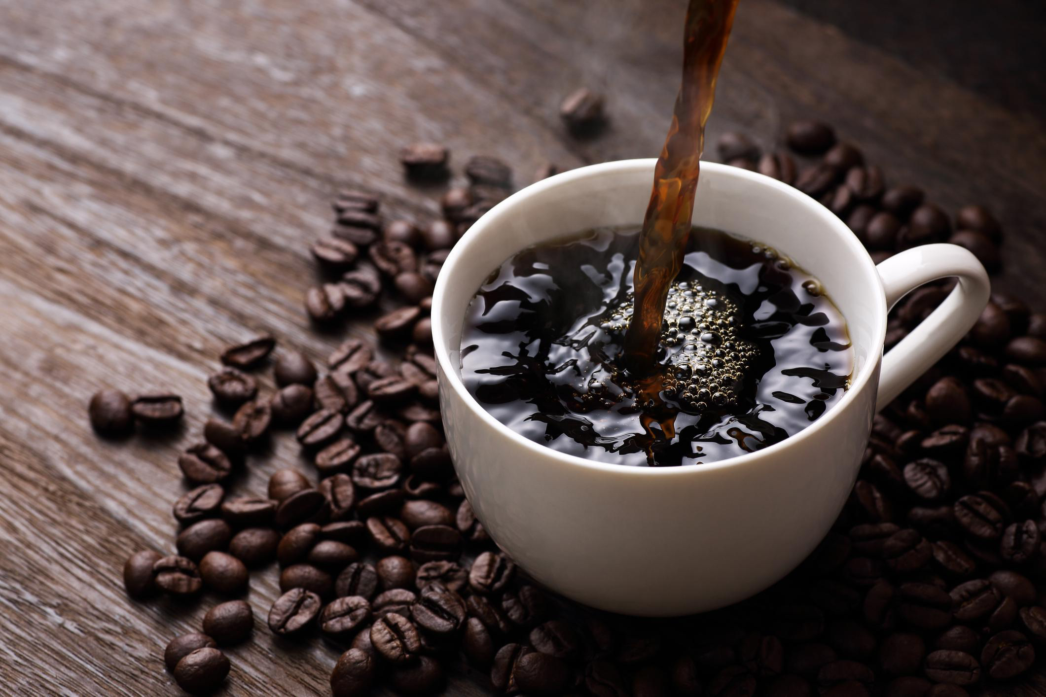 A cup of coffee being poured with coffee beans scattered around