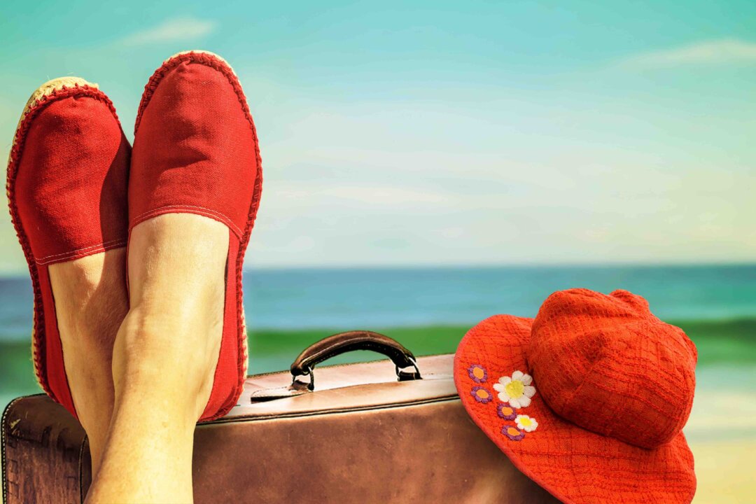 Legs with red cloth shoes in front of beach with a suitcase an a retro hat
