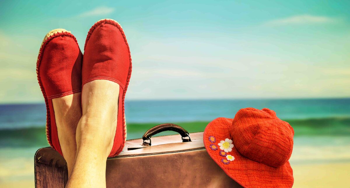 Legs with red cloth shoes in front of beach with a suitcase an a retro hat