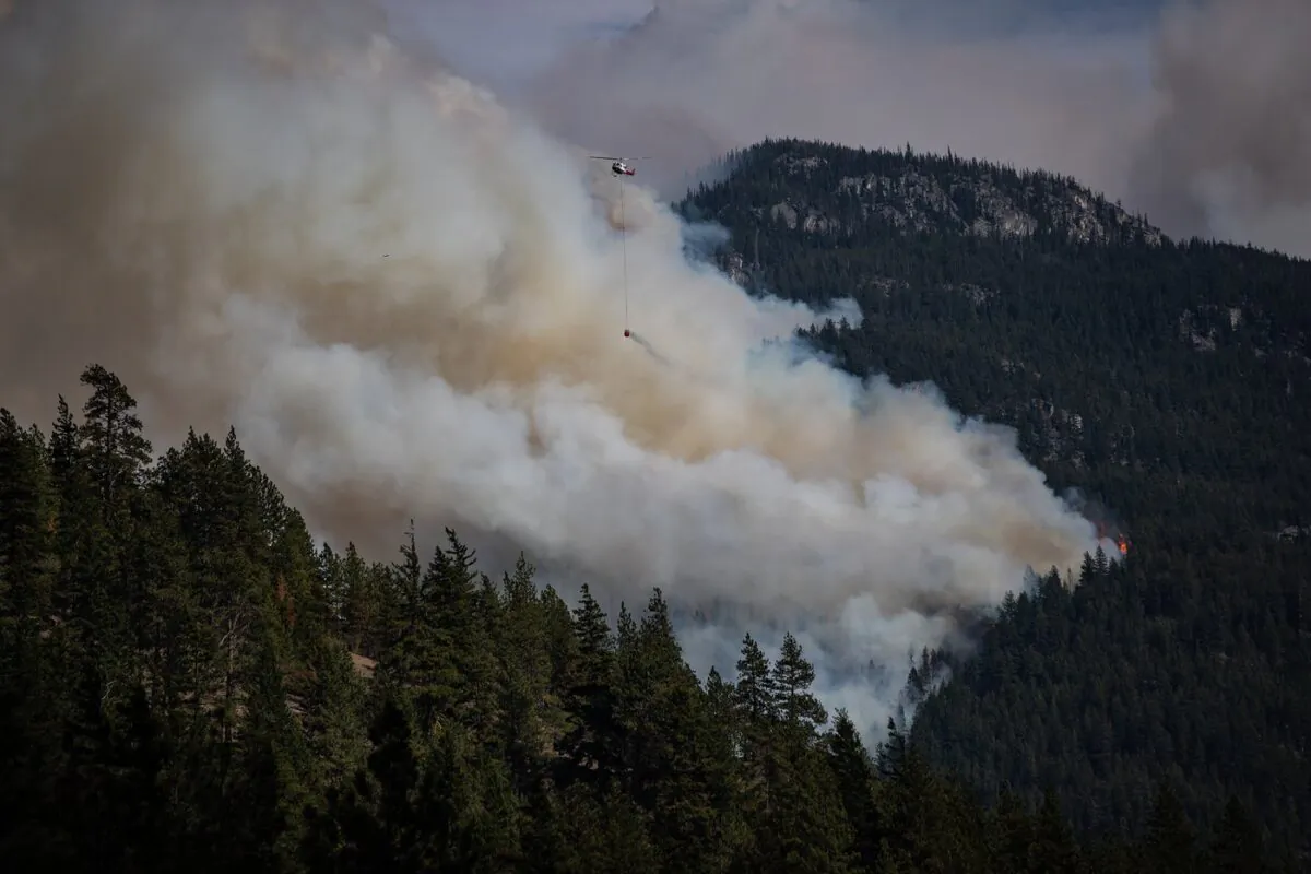A helicopter carrying a water bucket flies past the Lytton Creek wildfire burning in the mountains near Lytton, B.C., on Aug. 15, 2021. (The Canadian Press/Darryl Dyck)