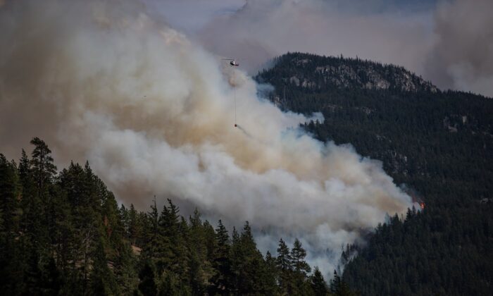 A helicopter carrying a water bucket flies past the Lytton Creek wildfire burning in the mountains near Lytton, B.C., on Aug. 15, 2021. (The Canadian Press/Darryl Dyck)