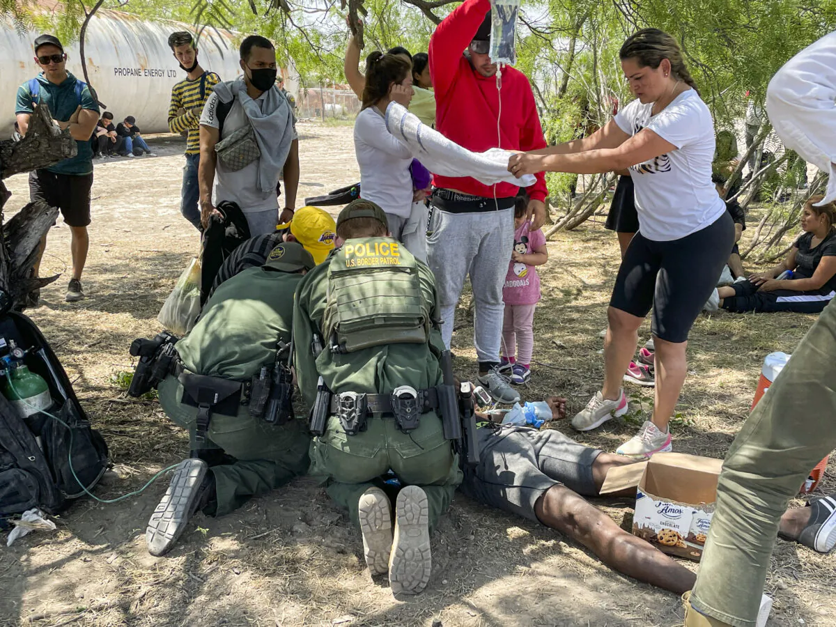 Border Patrol agents provide medical assistance to an illegal immigrant who has heat-related issues, while other agents apprehend a large group of illegal immigrants near Eagle Pass, Texas, on May 20, 2022. (Charlotte Cuthbertson/The Epoch Times)