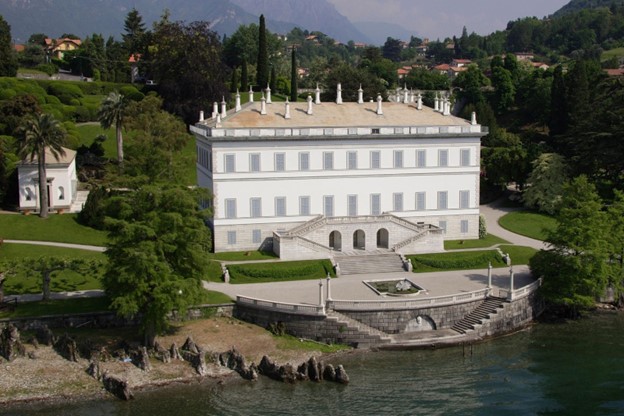 Villa Melzi has been declared a National Monument. The exterior double staircase rises up to the main floor and frames three arched doors that provide passage to the villa’s lower floor. The rusticated base and large textured brick-like forms establish a relationship with the ground, while providing contrast and hence highlighting the soft white walls. This treatment extends vertically along the corners, known as coins. The coins form a frame to the subdued façades comprised of a soft repetition of rectangular windows. The roof is adorned with chimney spires. (MT–Afb/Giardini di Villa Melzi)