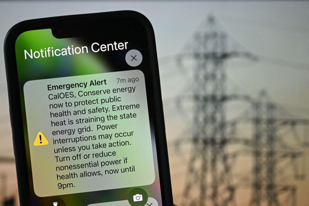 На фотоиллюстрации показан фон инфраструктуры электроснабжения с Apple iPhone, демонстрирующим уведомление о чрезвычайной ситуации от CalOES, призывающее население экономить энергию для защиты здоровья и безопасности, поскольку электросеть напряжена во время жары в Лос-Анджелесе, штат Калифорния, 6 сентября 2022 года. (Патрик Т. Фэллон / AFP через Getty Images)