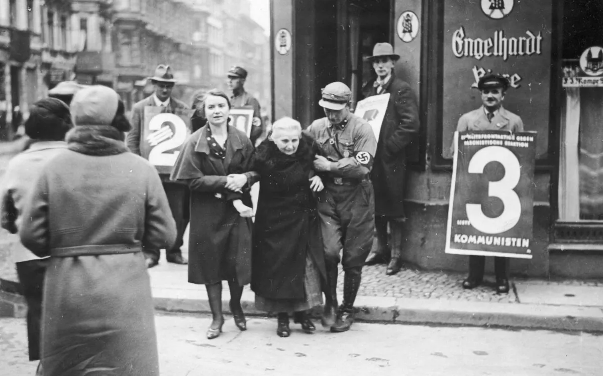 A Nazi Party member helps an elderly women from a polling station during the German federal election, Berlin, in November 1932. On the right is a man holding a placard for the Communist Party of Germany (KPD). (FPG/Archive Photos/Getty Images)