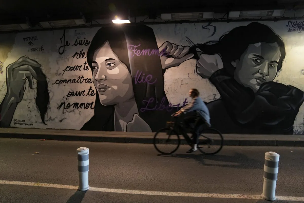 A woman rides bicycle front of a mural signed by Clacks-one and Heartcraft_Street art, depicting women cutting their hair to show support for Iranian protesters standing up to their leadership over the death of a young woman in police custody, in a tunnel in Paris, France, on Oct. 5, 2022. (Francois Mori/AP Photo)