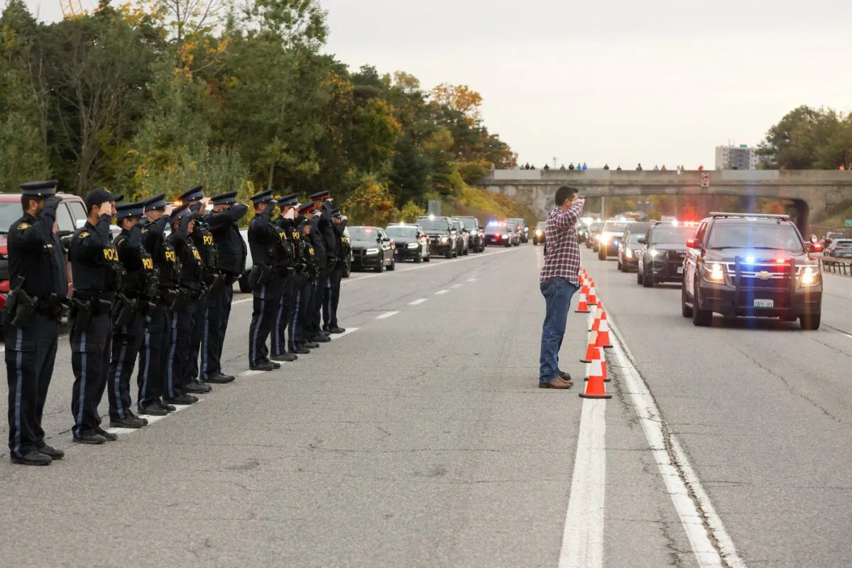 Ontario Provincial Police officer Adam McCourt, left in plainclothes, and Sgt. Jonathan Bouchard, obscured, salute the arriving procession carrying the bodies of two South Simcoe Police officers who were killed responding to a call, in Innisfil, Ont., Oct. 14, 2022. (The Canadian Press/Christopher Drost)