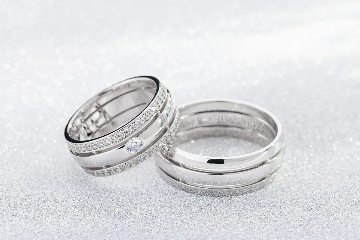 Pair of silver wedding rings with diamonds on white glossy background with bokeh. White gold wedding ring bands with gemstones on female ring. (TorriPhoto/Shutterstock)