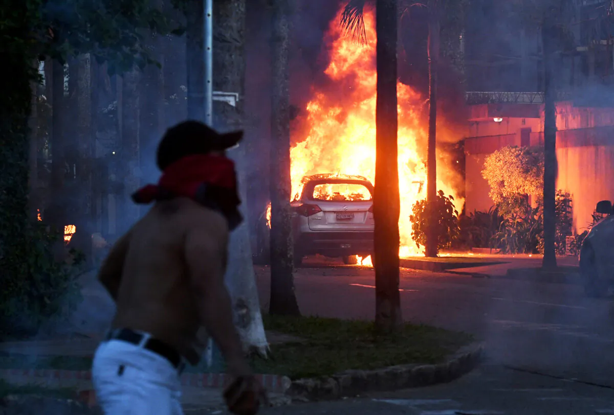 A vehicle burns as a demonstrator throws a stone at police during riots protesting the arrest of against the arrest of the Governor of Santa Cruz, Luis Fernando Camacho, in Bolivia, on Dec. 30, 2022. (Rodrigo Urzagasti/AFP via Getty Images)