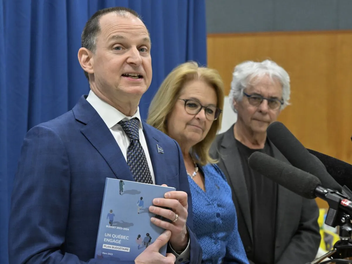 Quebec Finance Minister Eric Girard holds a copy of the 2023-24 budget speech as he responds to reporters at a news conference in Quebec City on March 20, 2023, Quebec Families Minister Suzanne Roy (C) and Dr. Gilles Julien look on. (The Canadian Press/Jacques Boissinot)