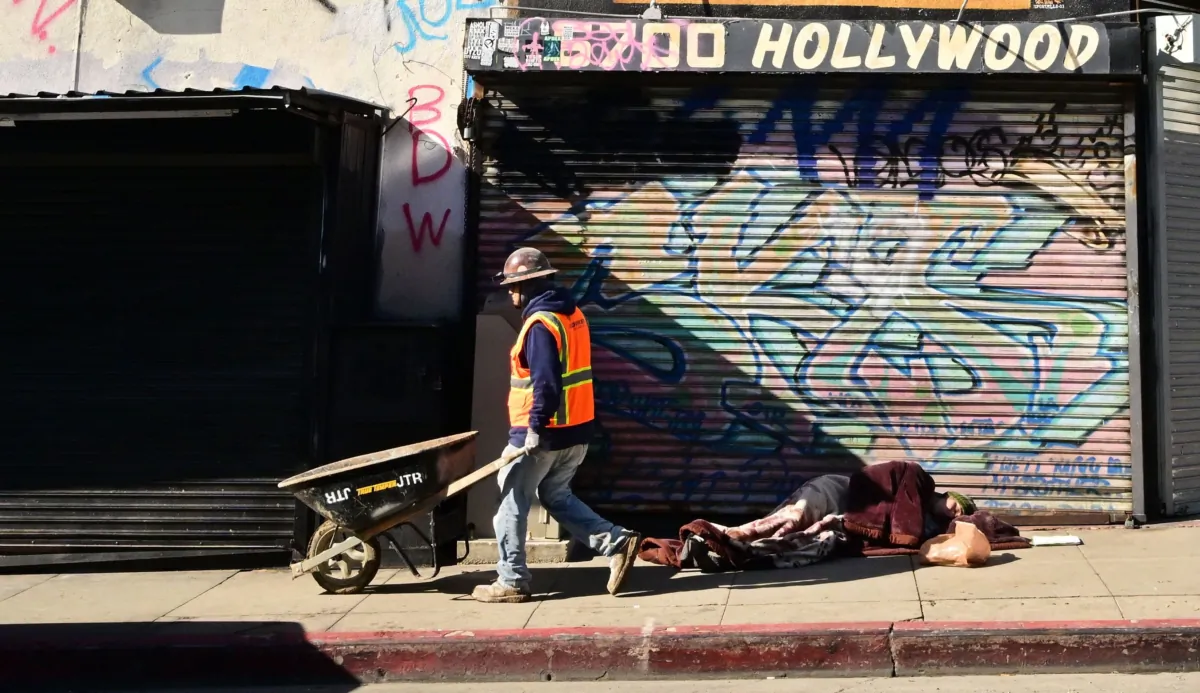 A construction worker pushes his wheelbarrow past a homeless man sleeping on a Hollywood sidewalk in Hollywood, Calif., on Jan. 20, 2023. (Frederic J. Brown/AFP via Getty Images)