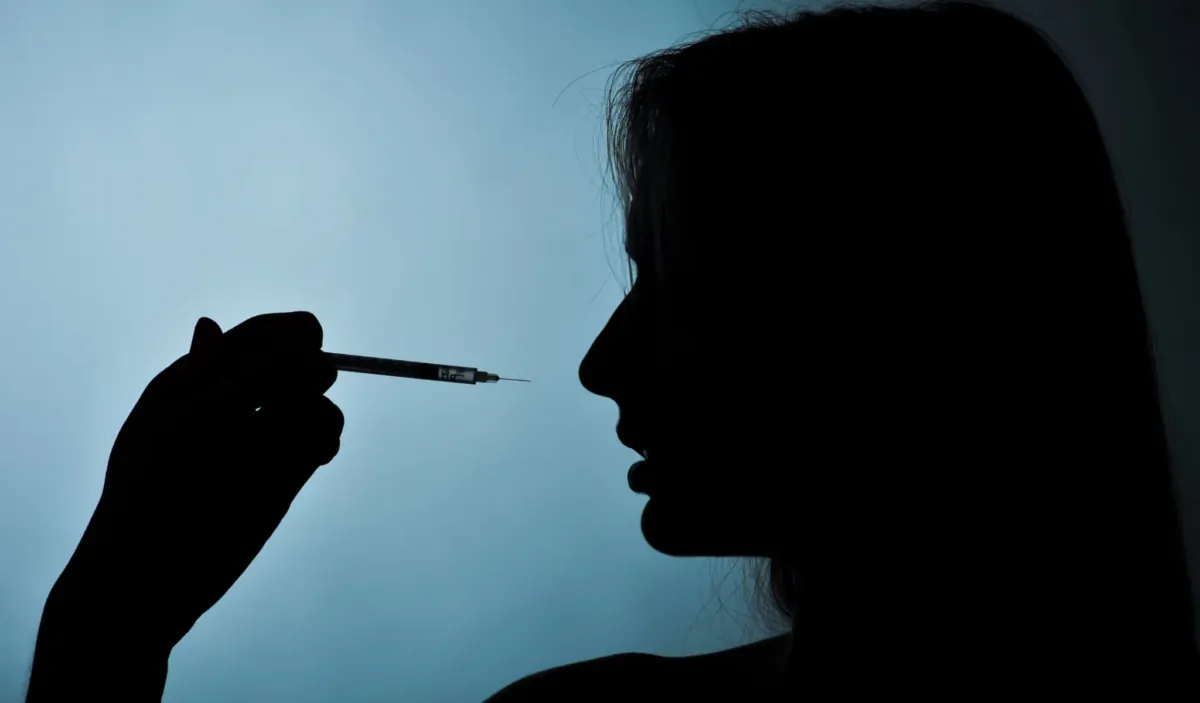 Syringe and drug or vaccine in hand and girl silhouette on a blue background. (Ruslan_127/Shutterstock)