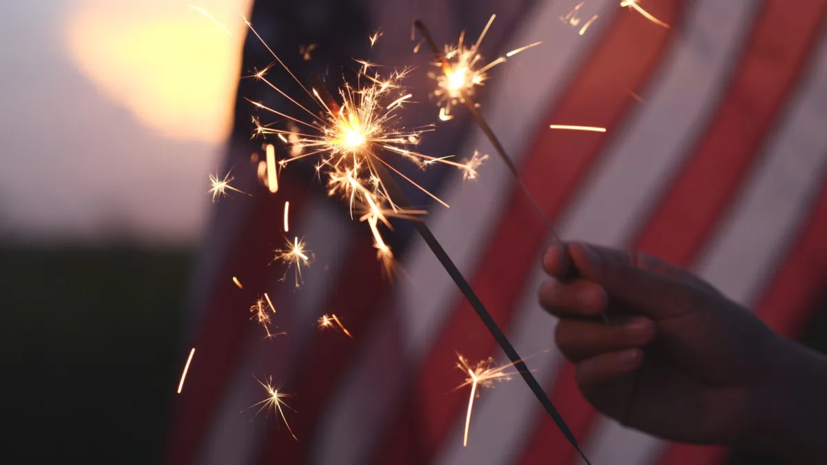 Stock photo of a hand holding sparkler fireworks with an American flag in the background. (iHumnoi/Shutterstock)