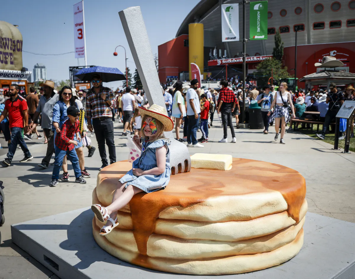 A young girl sits on a stack of pancakes for the 100th anniversary of the Stampede pancake breakfast on Family Day at the Calgary Stampede on July 9, 2023. (Jeff McIntosh/The Canadian Press)