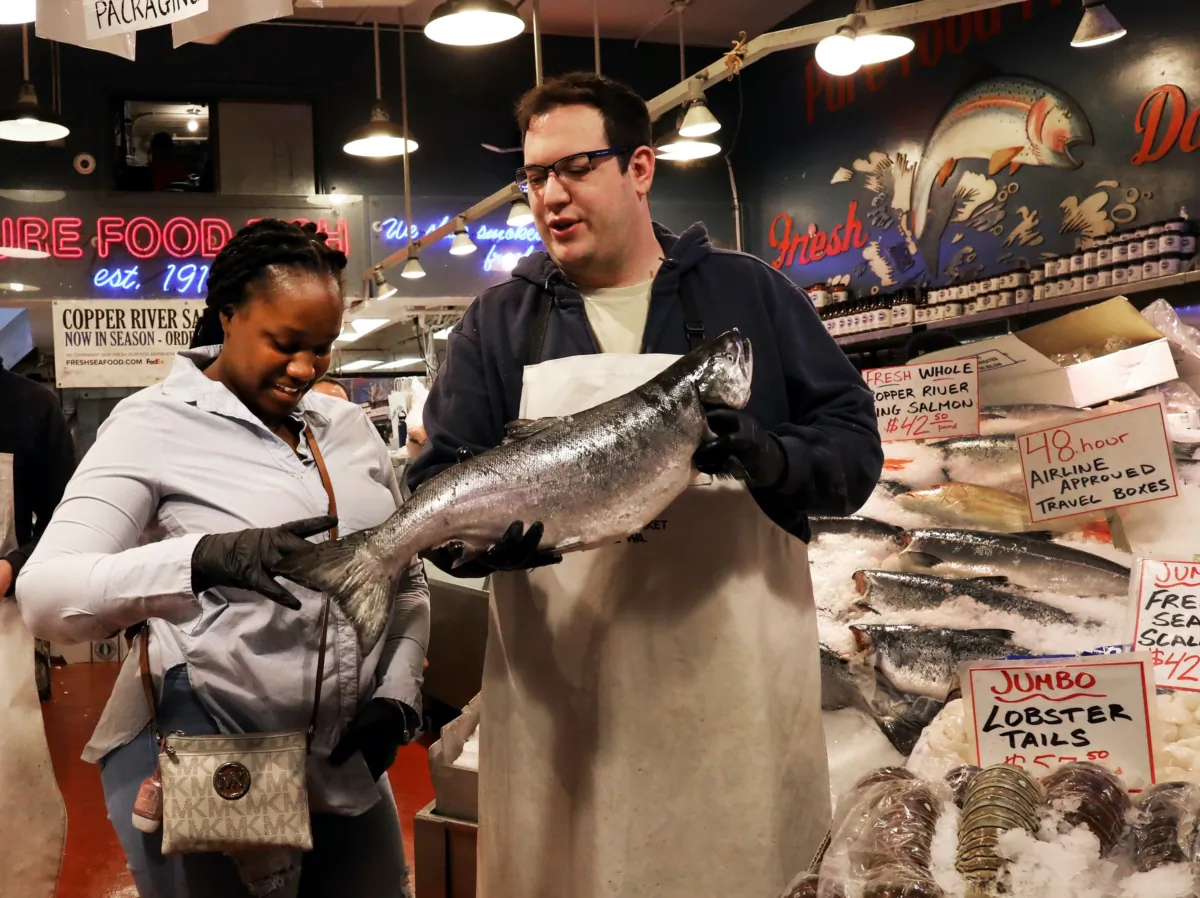 Joydie Ellis, visiting Seattle's Pike Place Market, wanted to have a souvenir photograph at the Pure Food Fish Market's stand and co-owner Isaac Behar suggested adding a king salmon to the image, on May 25, 2022. (Alan Berner/The Seattle Times/TNS)