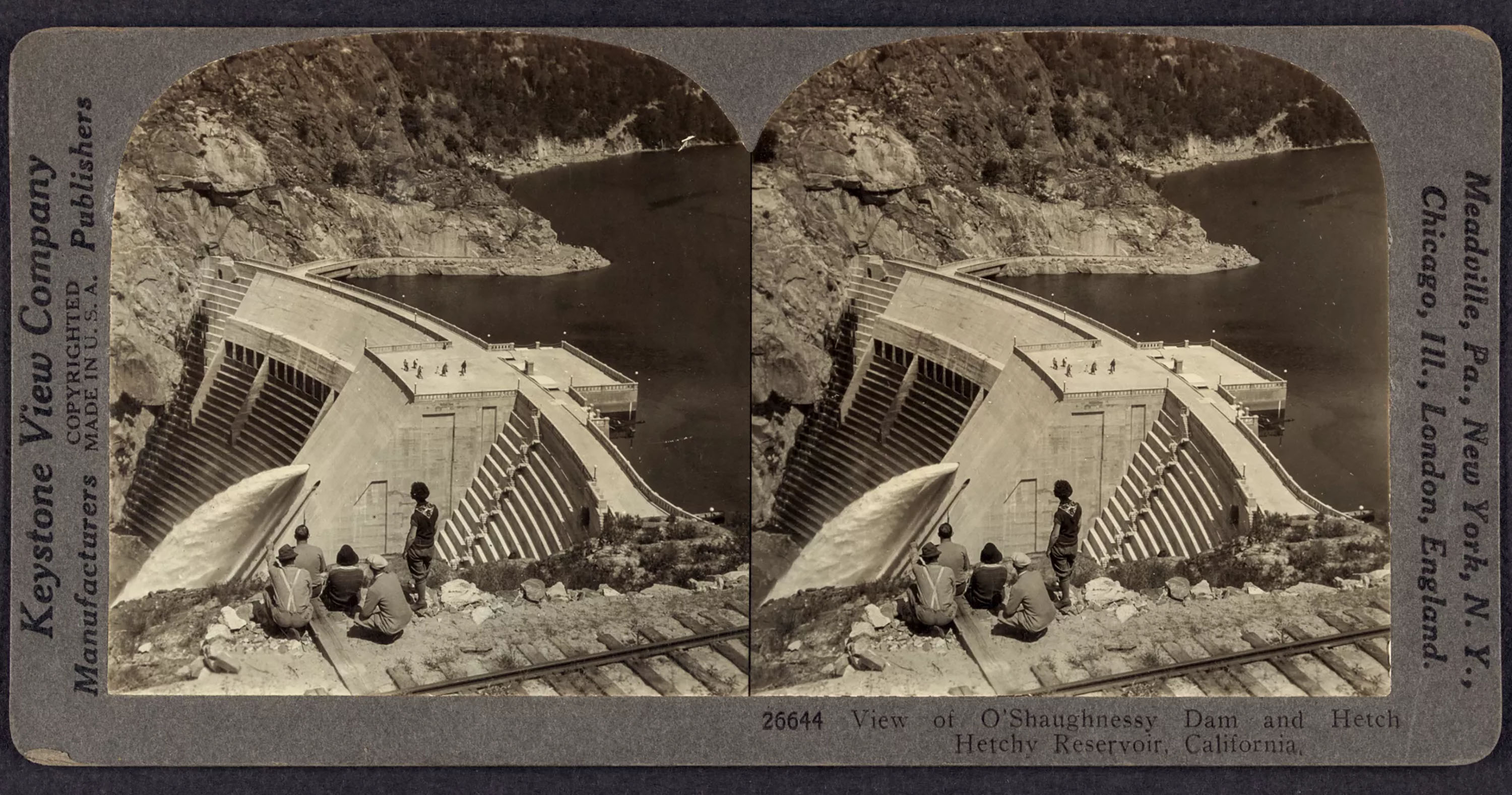 A stereograph from the Keystone View Company, circa 1926, showing a small group of people next to railroad tracks atop a hillside overlooking the O’Shaughnessy Dam and a portion of the Hetch Hetchy Reservoir in California. 