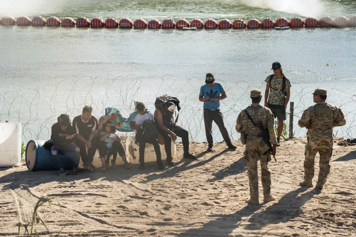 Asylum-seeking illegal immigrants sit by concertina wire fence while waiting to be transported by U.S. law enforcement officers after crossing the Rio Grande river into the U.S. from Mexico in Eagle Pass, Texas, on July 24, 2023. (Go Nakamura/Reuters)
