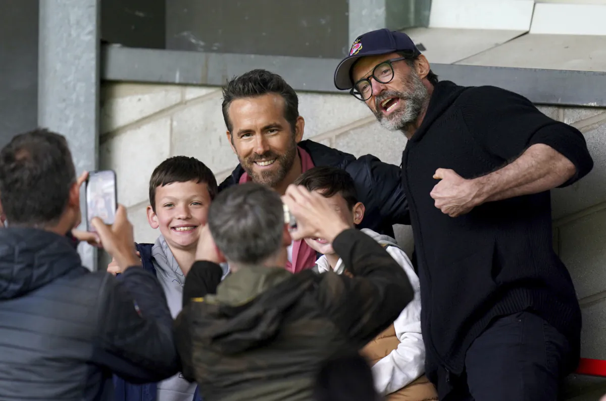 Wrexham Co-Owner, Ryan Reynolds, center, and Hugh Jackman, right, pose for a photo with a fans in the stands before the Sky Bet League Two soccer match between Wrexham and Milton Keynes Dons at the SToK Racecourse, Wrexham, Wales, on Aug. 5, 2023.  (Jacob King/PA via AP)