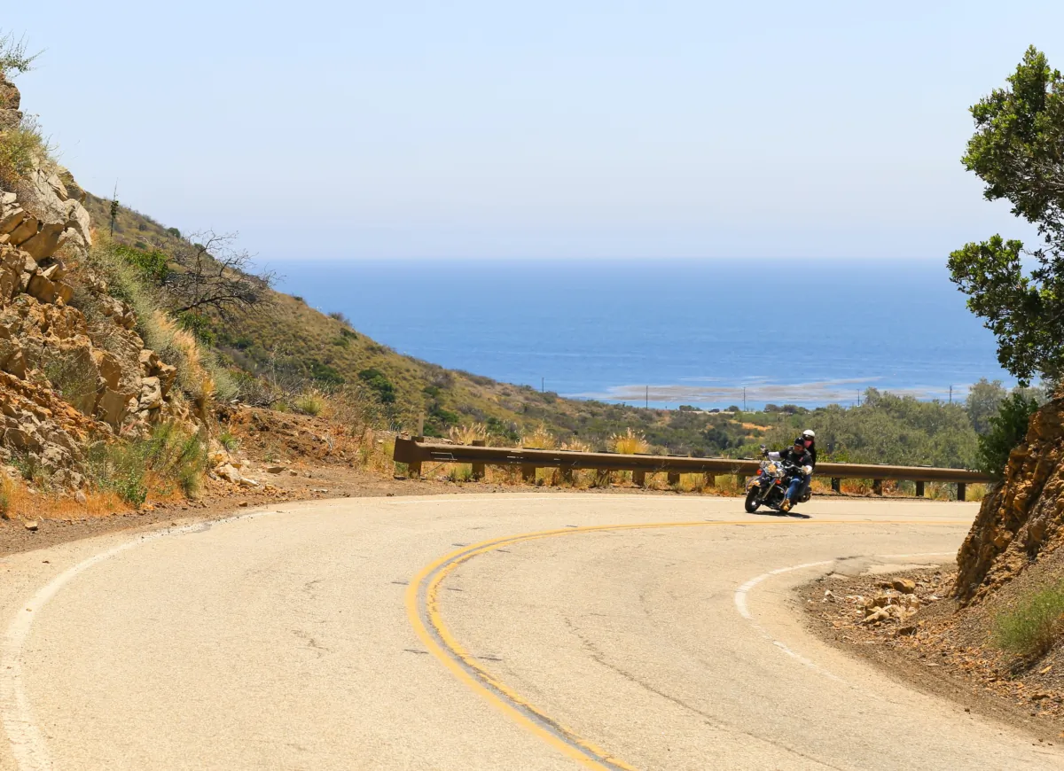 A biker and a pillion rider driving in a curve on the Mulholland Highway in the Malibu Hills. In the background is the Pacific Ocean. (Michael Rosebrock/Shutterstock)