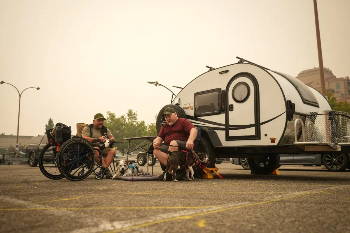 Veteran Rob Pullen, left, and Warren Pullen, who were evacuated from their home on Thursday due to wildfires, sit with their dogs outside their trailer in the parking lot at an evacuation centre, in Kelowna, B.C., on Aug. 19, 2023. (The Canadian Press/Darryl Dyck)