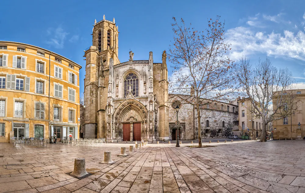 The west side of Aix Cathedral features a Gothic façade with intricate carvings and ornate details. The exterior combines various architectural styles, with Romanesque stone walls and flat square yellowish buttresses topped with pinnacles, contrasting with the central structure of the portal. (Sergey Dzyuba/Shutterstock)
