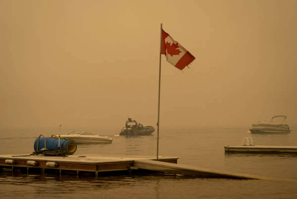 Thick smoke from the Lower East Adams Lake wildfire fills the air and a Canadian flag flies in the wind as RCMP officers on a boat patrol Shuswap Lake, in Scotch Creek, B.C., on Aug. 20, 2023. (The Canadian Press/Darryl Dyck)