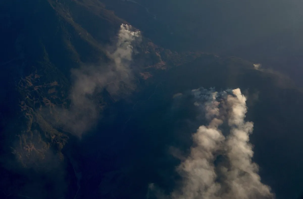 Smoke from wildfires can be seen in a mountain range from the passenger window aboard a charter flight between Penticton and Vancouver, on Aug. 22, 2023. (The Canadian Press/Chad Hipolito)