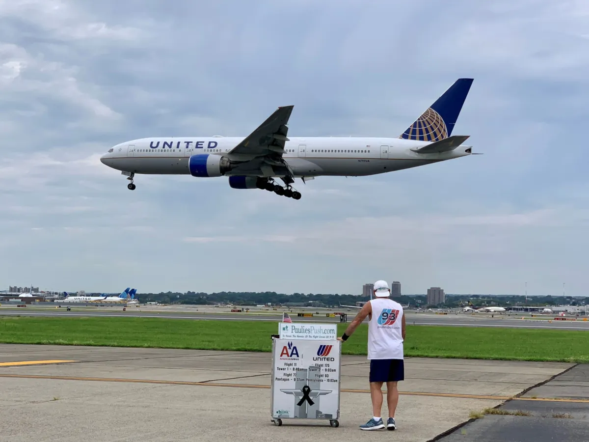An airplane flying over Paul “Paulie” Veneto, a retired flight attendant, as he pushes an airline beverage cart at Newark-Liberty International Airport in remembrance of the flight crews who died on 9/11. In Newark, N.J., on Aug. 14, 2023. (Courtesy Paulie’s Push) 