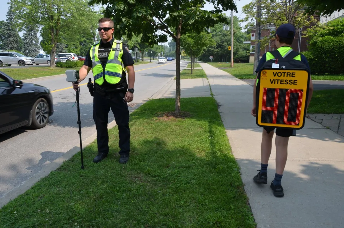 A police officer stands by as a child with a backpack, equipped with a readout showing drivers' speeds, in a June 30, 2023, photo. (The Canadian Press/HO-Service de police de l'agglomeration de Longueuil,)