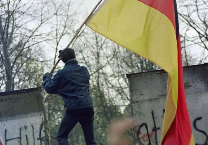 A West Berliner carries a Federal Republic of Germany flag to hand to an East German through a portion of the fallen Berlin Wall near the Brandenburg Gate on Nov. 11, 1989. Three leaders were instrumental in the fall of the Berlin Wall and the end of the Cold War: U.S. President Ronald Reagan, English Prime Minister Margaret Thatcher, and Polish Pope John Paul II. (Gerard Malie/AFP via Getty Images)